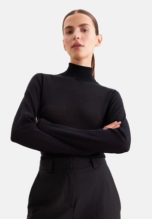 Jeune femme aux cheveux raides tirés en arrière, portant un pull à col roulé noir et un pantalon noir, bras croisés, sur un fond clair uni.