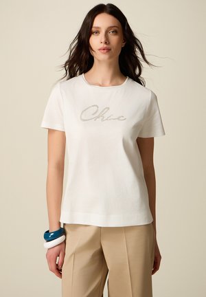 Donna con lunghi capelli scuri che indossa una t-shirt bianca decorata "Chloe", pantaloni beige e braccialetti blu e bianchi al polso sinistro.