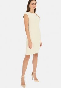 Dopasowana kremowa sukienka z krótkimi rękawami, okrągłym dekoltem i ozdobnymi detalami na ramionach. Noszona z cielistymi szpilkami.