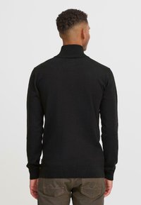 Une personne se tient de dos par rapport à la caméra, portant un pull à col roulé noir et un jean marron, sur un fond blanc uni.