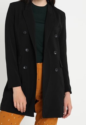 Femme portant un blazer croisé noir, un haut vert foncé et un pantalon moutarde avec un petit motif dispersé.