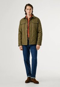 Casaco verde-oliva acolchoado com dois bolsos no peito e botões de pressão, usado sobre uma camisa cor de ferrugem, combinado com jeans azul-escuro e botas castanhas.