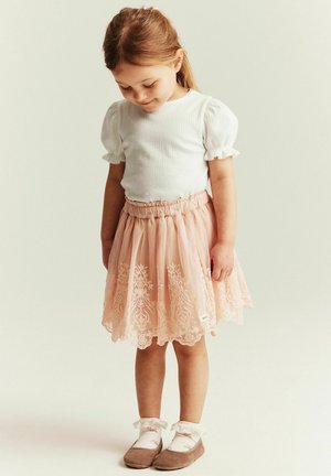 Jeune fille aux cheveux châtain clair regardant vers le bas, portant un haut blanc à manches bouffantes, une jupe en dentelle rose, des chaussettes fleuries et des chaussures marron.