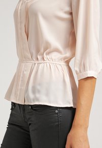 Jasnoróżowa, jedwabna bluzka z rękawami o długości 3/4, marszczonym pasem i pionowymi fałdami na przodzie. Połączona z czarnymi spodniami o dopasowanym kroju.