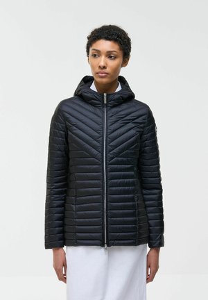 Donna che indossa una giacca imbottita nera con cappuccio chiusa con la cerniera, in piedi contro uno sfondo chiaro uniforme, con le mani rilassate lungo i fianchi.