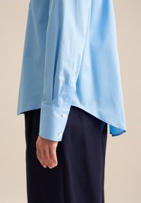 Camicia azzurra a maniche lunghe con orlo arrotondato, polsini con bottoni e tessuto di cotone morbido. Indossata sopra pantaloni scuri, che evidenziano una vestibilità rilassata.