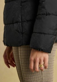 Parem käsi, millel on kaks kuldringi, on asetatud houndstooth mustriga pükste ja musta puhvjakki varruka kõrvale, millel on "Joules" logo beežil taustal.