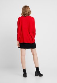 Roter Strickpullover mit lockerem Schnitt, gerippter Struktur und langen Ärmeln, kombiniert mit einem schwarzen Rock und Stiefeletten.