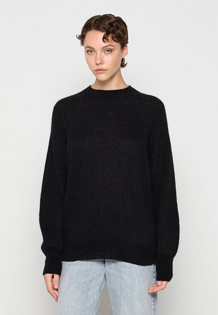 Schwarzer Strickpullover mit Rundhalsausschnitt, langen Ärmeln und lockerer Passform. Gerippte Textur und ein leicht verlängeter Saum. Kombiniert mit hellblauen Jeans.