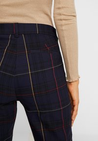 Pantalons à carreaux navy avec des lignes rouges et dorées, dotés de deux poches arrière et d'une taille haute, associés à un haut beige à manches longues.