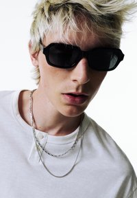 Ochelari de soare hexagonali negri, ten deschis, purtând o cămașă henley albă. Două lanțuri din argint: unul plat, unul rotund, așezate pe guler.