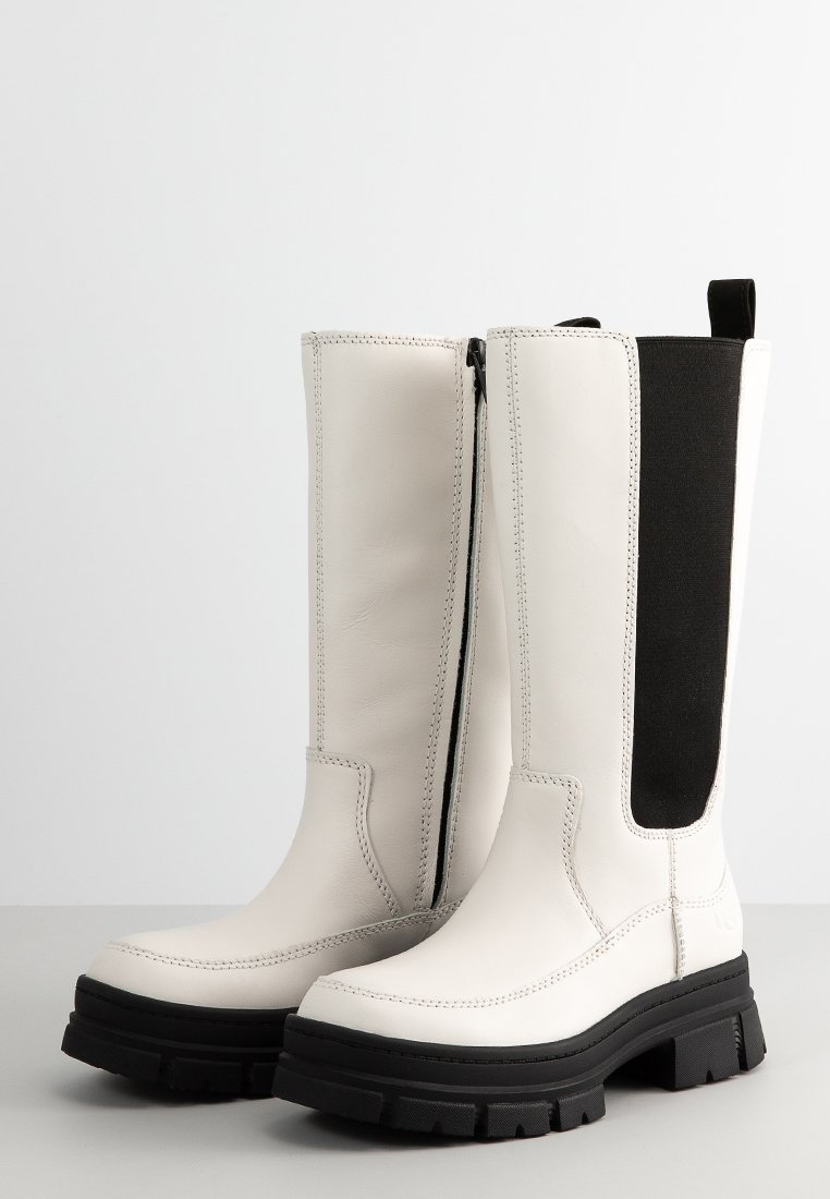 Weiße Lederstiefel bis zum Knie haben ein schwarzes elastisches Panel, einen seitlichen Reißverschluss, eine klobige schwarze Gummisohle und sichtbare Nähdetails.