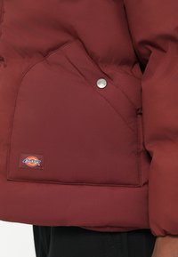 Maroon puffer jakna s dijagonalnom džepom s kopčanjem na dugme i logotipom na rubu. Glatka, izdržljiva tekstura tkanine.