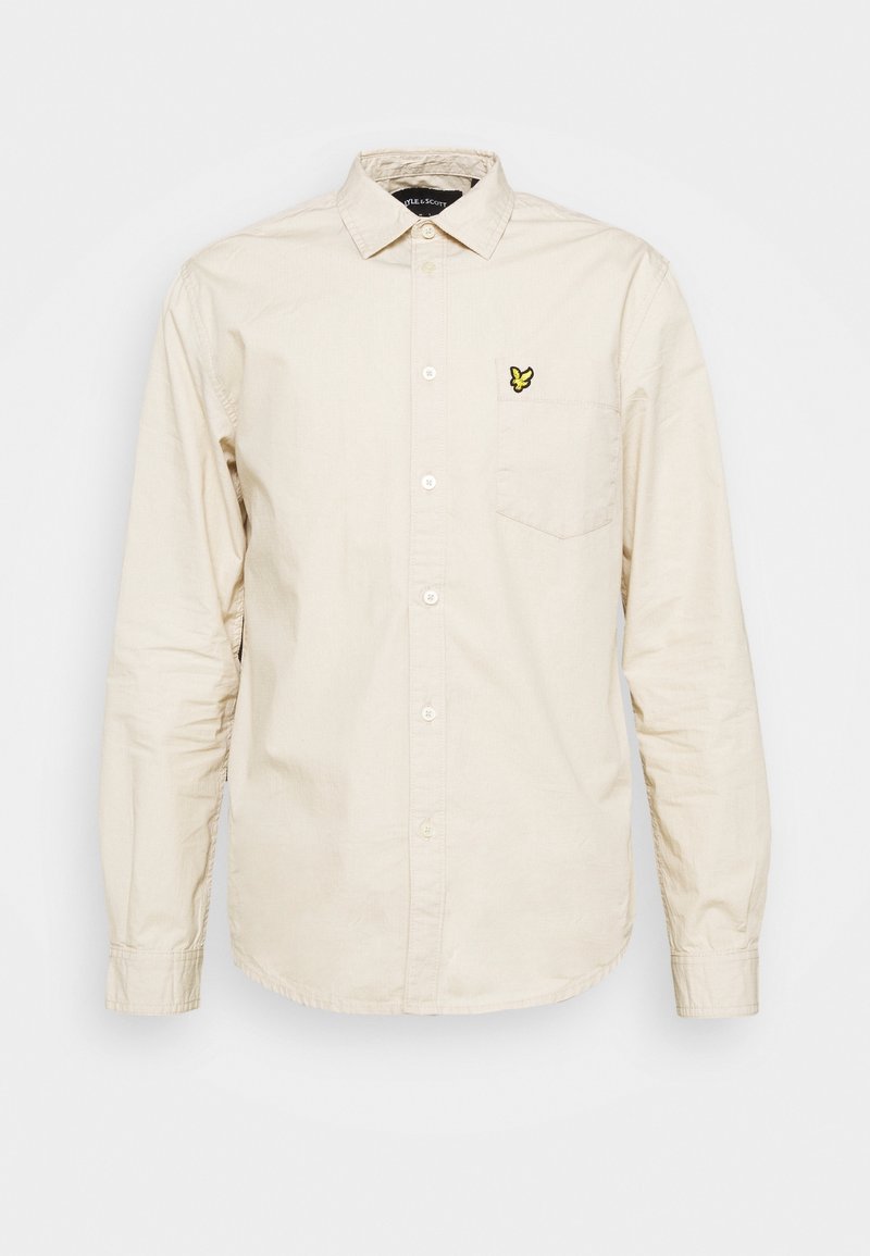 Beige långärmad skjorta med knappar, krage, bröstficka och liten gul fågel-logotyp på fickan.