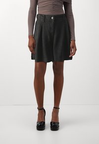 Zwarte geplooide shorts van een zachte stof met een knoopsluiting. Draagt een strakke top met lange mouwen en glanzende zwarte platformhakken.