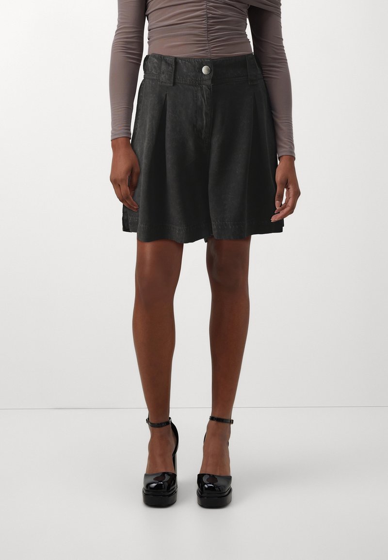 Zwarte geplooide shorts van een zachte stof met een knoopsluiting. Draagt een strakke top met lange mouwen en glanzende zwarte platformhakken.