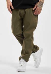 Pantaloni in cotone verde oliva con una vestibilità rilassata, con orlo alla caviglia, abbinati a sneakers bianche, dotati di cuciture visibili e tasche laterali.