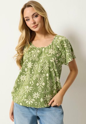 Frau mit langen blonden Haaren, die eine grüne gemusterte Kurzarmbluse und hellblaue Jeans trägt, steht vor einem einfarbigen hellen Hintergrund.