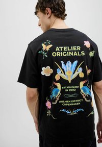 Černé bavlněné tričko s barevnými květinovými a ptákovými grafikami na zadní straně, se textem "ATELIER ORIGINALS" a detaily o lokalitě.