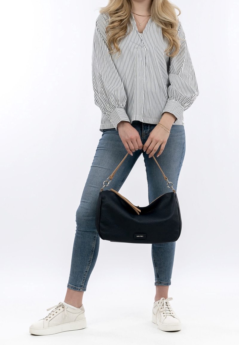 Femme portant un chemisier rayé et un jean bleu, tenant un sac à main noir avec une sangle beige, debout devant un fond blanc.