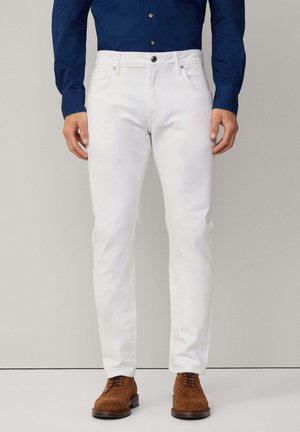 White denim jeans with a slim fit, featuring five pockets, a front button closure, and subtle stitching details, paired with brown suede shoes.
