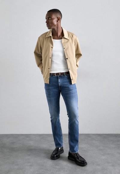 Beez värvi nööpidega jakk, mis on valge nahktagi peal, koos helesiniste denim-teksade ja mustade kingadega. Sile kangas, mugav lõige, kaasaegne disain.