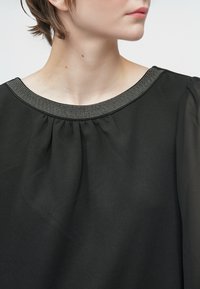 Woman wearing black blouse with sheer sleeves and beaded neckline, head turned to the right against a plain white background.
