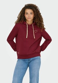 Bordeaux hættebluse med beige snore og hjertebroderi på brystet, lavet af et blødt stof, parret med lyse jeans.