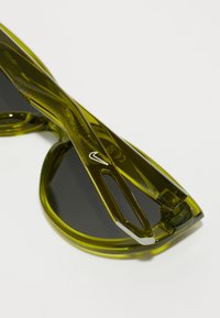 Ochelari de soare verzi, cu un cadru texturat, formă elegantă și lentile negre. Prezentând detalii gravate ale brandului pe braț și o finisare netedă.