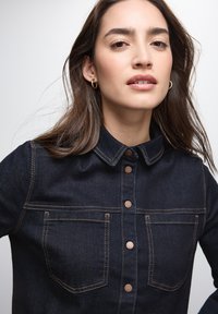 Giubbotto in denim scuro con colletto, chiusura a bottoni e due tasche sul petto. Presenta cuciture a contrasto e bottoni in bronzo.