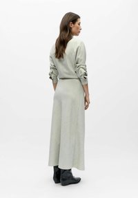 Robe vert clair texturée à manches longues et corsage cintré, s'évasant en une jupe ample, associée à des bottines noires.