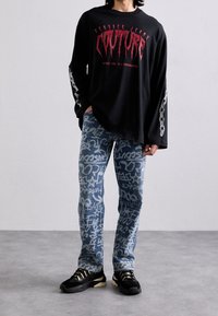 Musta pitkähihaisen t-paita, jossa on punainen graafinen teksti, yhdistettynä vaaleansinisiin farkkuihin, joissa on valkoisia graffitikuoseja. Mustat lenkkitossut kultaisilla yksityiskohdilla.