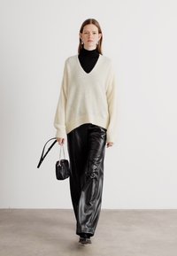 Maglione oversize beige chiaro con scollo a V, abbinato a pantaloni in pelle neri, larghi. La modella porta una piccola borsa nera con tracolla a catena.