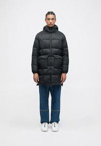 Giovane in piedi, con un cappotto imbottito nero, jeans blu e sneaker bianche, su uno sfondo chiaro e semplice.