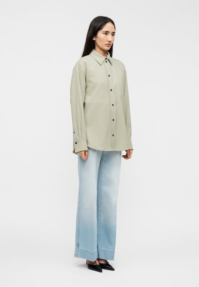 Φόρεμα σε ανοιχτό πράσινο χρώμα, υπερμεγέθη πουκάμισο με κλείσιμο με κουμπιά, μακριά μανίκια και κανονικό γιακά· συνδυασμένο με ανοιχτό μπλε, φαρδιά τζιν παντελόνια και μαύρες γόβες.