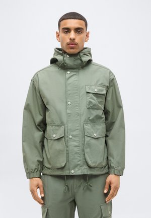 Homme portant une veste à capuche vert olive avec des poches à boutons-pression et un pantalon assorti sur un fond uni.