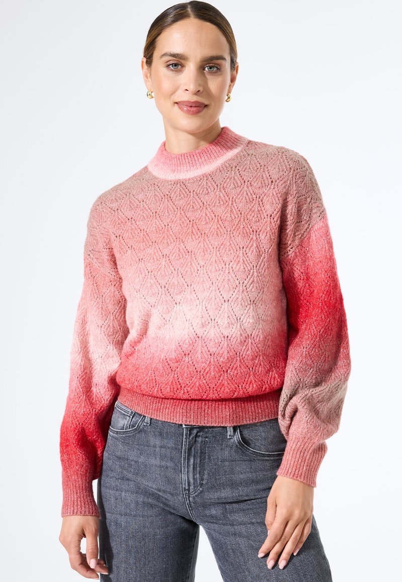 Gebreide trui met een verloop van lichtroze naar rood, met een getextureerd diamanten patroon en geribde boorden en zoom. Gepaard met grijze jeans.