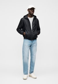 Μαύρο ελαφρύ μπουφάν με κουκούλα, γκρι μπλουζάκι, ανοιχτό μπλε τζιν και λευκά αθλητικά παπούτσια. Απλός σχεδιασμός, ομαλή υφή και casual εφαρμογή.