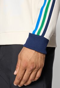 Nærbilde av en hånd som hviler på siden, iført en kremfarget sweatshirt med blå, grønn og marineblå striper på ermekanten.
