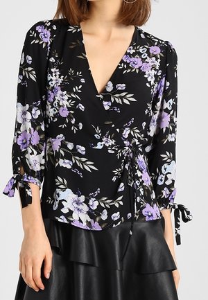 Mujer con blusa negra cruzada con estampado floral en morado y blanco y mangas tres cuartos con detalles de lazos, combinada con una falda de cuero negra.