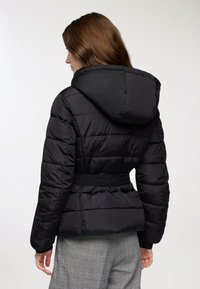 Czarna kurtka puchowa z kapturem, z wąskim wcięciem w talii, poziomymi szwami i gładką teksturą. Nosi na sobie spodnie w kratę.