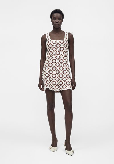 Γυναίκα στέκεται φορώντας ένα λευκό και καφέ φλοράλ μίνι φόρεμα από κροσέ χωρίς μανίκια και άσπρα μυτερά τακούνια με λεπτομέρειες φιόγκου.