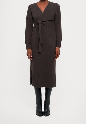 Femme portant une robe midi à manches longues marron foncé avec un détail noué à l'avant et des bottes noires montantes jusqu'aux genoux, debout devant un fond blanc.