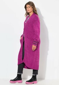 Lange, open voorkant cardigan in levendig magenta breiwerk, met geribbelde manchetten en twee voorzakken, gecombineerd met zwarte jeans en laarzen met een roze zool.