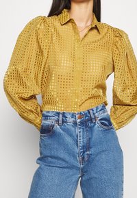 Blouse à carreaux jaune moutarde avec manches bouffantes, en tissu transparent, associée à un jean bleu taille haute avec coutures visibles.