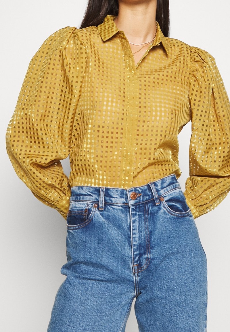 Blouse à carreaux jaune moutarde avec manches bouffantes, en tissu transparent, associée à un jean bleu taille haute avec coutures visibles.