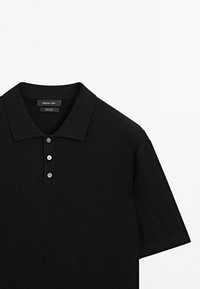 Svart ribbad polo med korta ärmar, tre knappslå och en platt krage. Tillverkad av mjuk bomull med en texturerad yta.