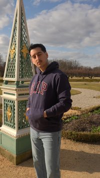 Jonge man met handen in de zakken, gekleed in een marineblauwe GAP-hoodie en lichte jeans, staand bij een decoratieve groene en gouden obelisk in een tuin.