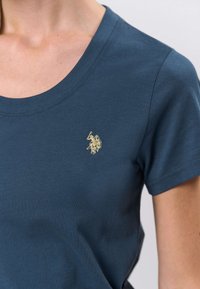 Tamnoplava majica kratkih rukava s okruglim izrezom i zlatnim vezenim logotipom na lijevoj strani prsa. Mekana tkanina s glatkom teksturom.