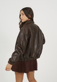 Brązowa skórzana kurtka bomberka o luźnym kroju, z elastycznym dołem, dużym kołnierzem i zaokrąglonymi ramionami, noszona na plisowanej spódnicy.